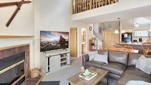 A modern living room with a fireplace, a large flat-screen TV, a sofa, and a dining area with wooden furniture. - Woods  Resort Village 10 Cozy Killington Escape –1BR + Loft, Fireplace, Pool & Hot Tub Access (Killington)