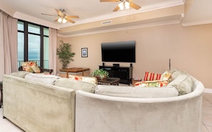 A living room with a large L-shaped sectional sofa, a flat-screen TV, a ceiling fan, and a large window with curtains.