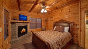 A log cabin bedroom with a stone fireplace, a bed with a headboard, and a ceiling fan. - Serenity Lodge: Theater, Gameroom, Hot Tub (Gatlinburg)