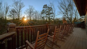 A wooden deck with rocking chairs overlooking a forested area during sunset. - Serenity Lodge: Theater, Gameroom, Hot Tub (Gatlinburg)