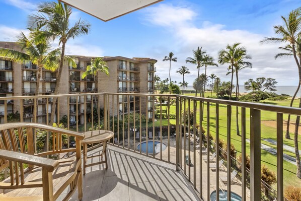 A balcony with a wooden deck, a pool, and a view of a building with multiple balconies and palm trees.