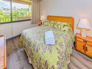 A hotel room with a bed, two bedside lamps, a wooden nightstand, and a view of greenery outside. - West Maui Suite Life w/Island Décor, Kitchen+Lanai, WiFi–Kamaole Sands 8302 (KIHEI)