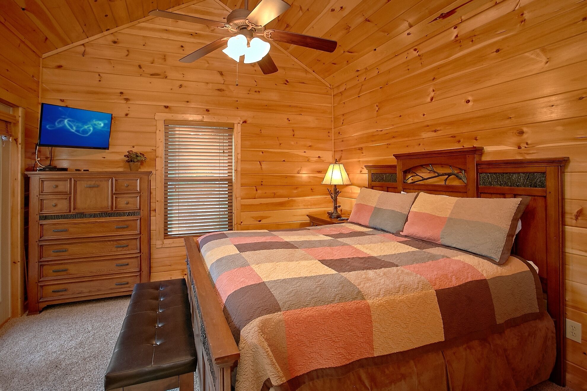 Ein gemütliches Schlafzimmer in einer Holzhütte mit Bett, Bank, Kommode, Fernseher und Deckenventilator.