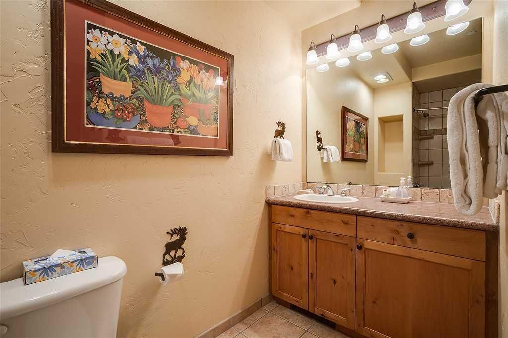 Une salle de bain avec des meubles en bois, un lavabo, des toilettes, un tableau floral encadré et un espace douche.