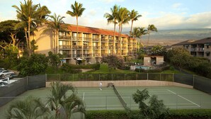 Un campo da tennis circondato da lussureggiante vegetazione e palme, con un edificio di un hotel sullo sfondo.