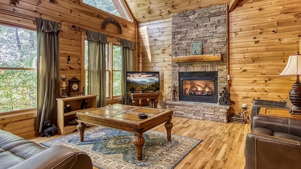 A cozy cabin interior with wooden walls, a stone fireplace, a wooden table, and a leather couch. - Whispering Oaks - Private Pigeon Forge Getaway...Close to Tanger Outlet Shopping (Sevierville)