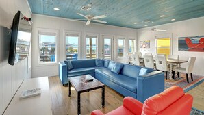 A modern living room with a blue sectional sofa, a red armchair, a wooden coffee table, and a dining area with white chairs and a table. - Come stay and play at the Cherry on Top, Ocean Village’s premier rental! Private (Port Aransas)