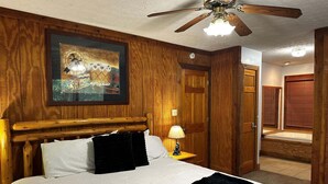 Ein Zimmer mit Holzwänden, einem Bett, einem Deckenventilator, einem Gemälde und einer Tür zu einem anderen Raum.