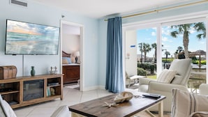 Un salon moderne comprenant une télévision, une table basse en bois, un canapé et un balcon offrant une vue sur des palmiers et une piscine.