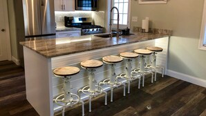 A modern kitchen with stainless steel appliances, a central island with bar stools, and a window with blinds. - Renovated Cozy Chic North Fork Cottage, steps away from the Beach & Wine Trail!! (Calverton)