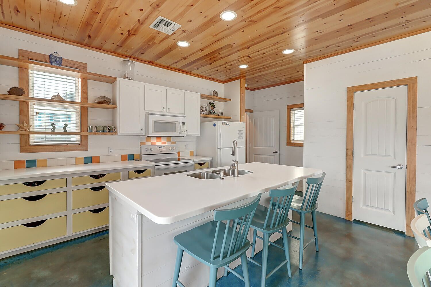 A kitchen with white cabinets, a microwave, a stove, and a sink. There are two blue bar stools and a window with blinds.
