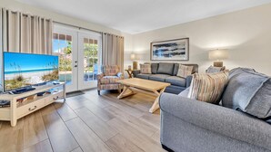 A living room with a TV, sofa, and armchair, a wooden coffee table, and a view of the beach through a glass door. - Beach Service Included, Private Beach, Chateau La Mer 9B! (Destin)