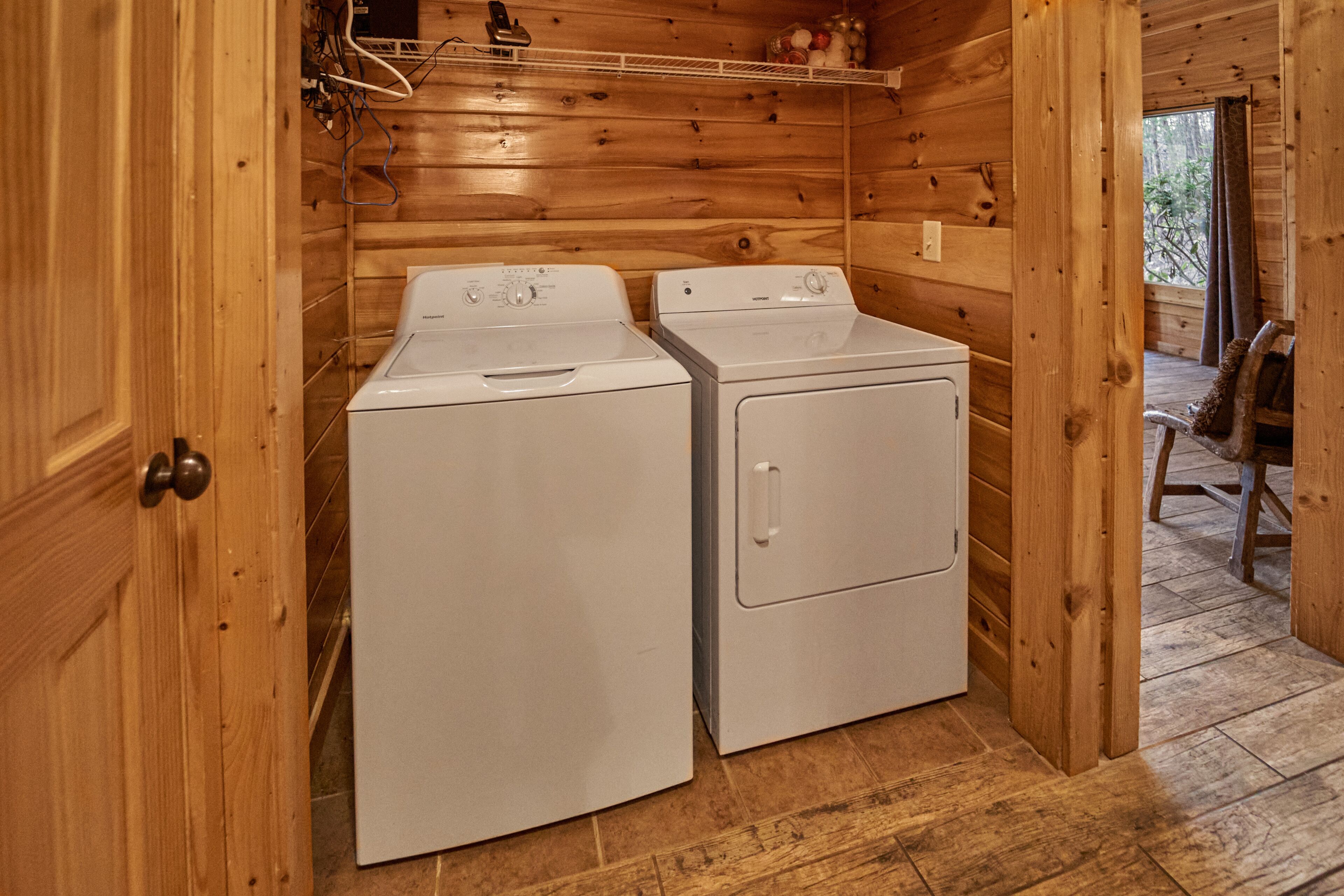 A laundry room with two white washers and dryers, wooden walls, and a window with a view of trees.