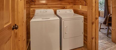 A laundry room with two white washers and dryers, wooden walls, and a window with a view of trees.
