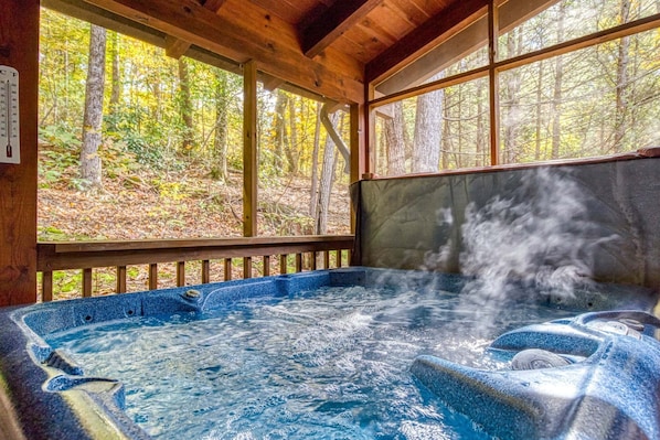 A hot tub with steam rising, surrounded by wooden deck and large windows overlooking a forest.