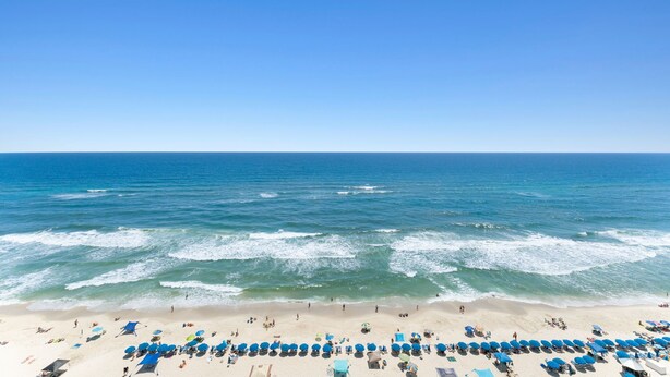 A beach with rows of blue umbrellas and beach chairs, a sandy shore, and the ocean. - Beachy, Relaxing, Beachfront Condo, Great Location, Awesome Balcony Features (Panama City Beach)