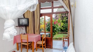 Une pièce avec une table et des chaises en bois, un téléviseur posé sur un meuble, et une vue sur un jardin par une porte vitrée.