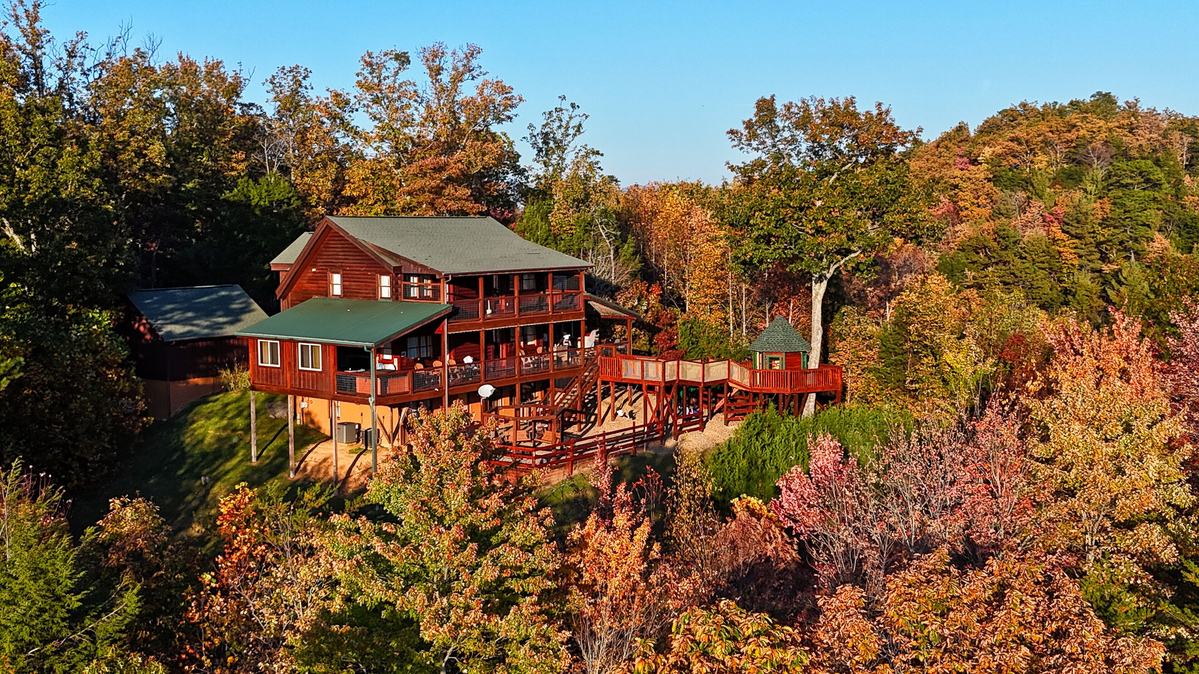A large wooden cabin with multiple decks and a covered porch, surrounded by dense forest and autumn foliage.