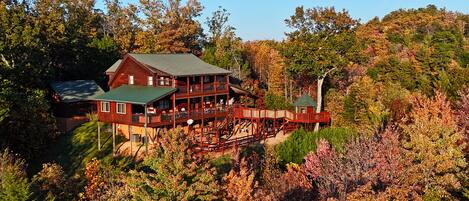 A large wooden cabin with multiple decks and a covered porch, surrounded by dense forest and autumn foliage.