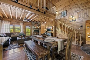 Interior de una cabaña de madera con comedor, escalera y ventanas amplias que ofrecen vistas al exterior.