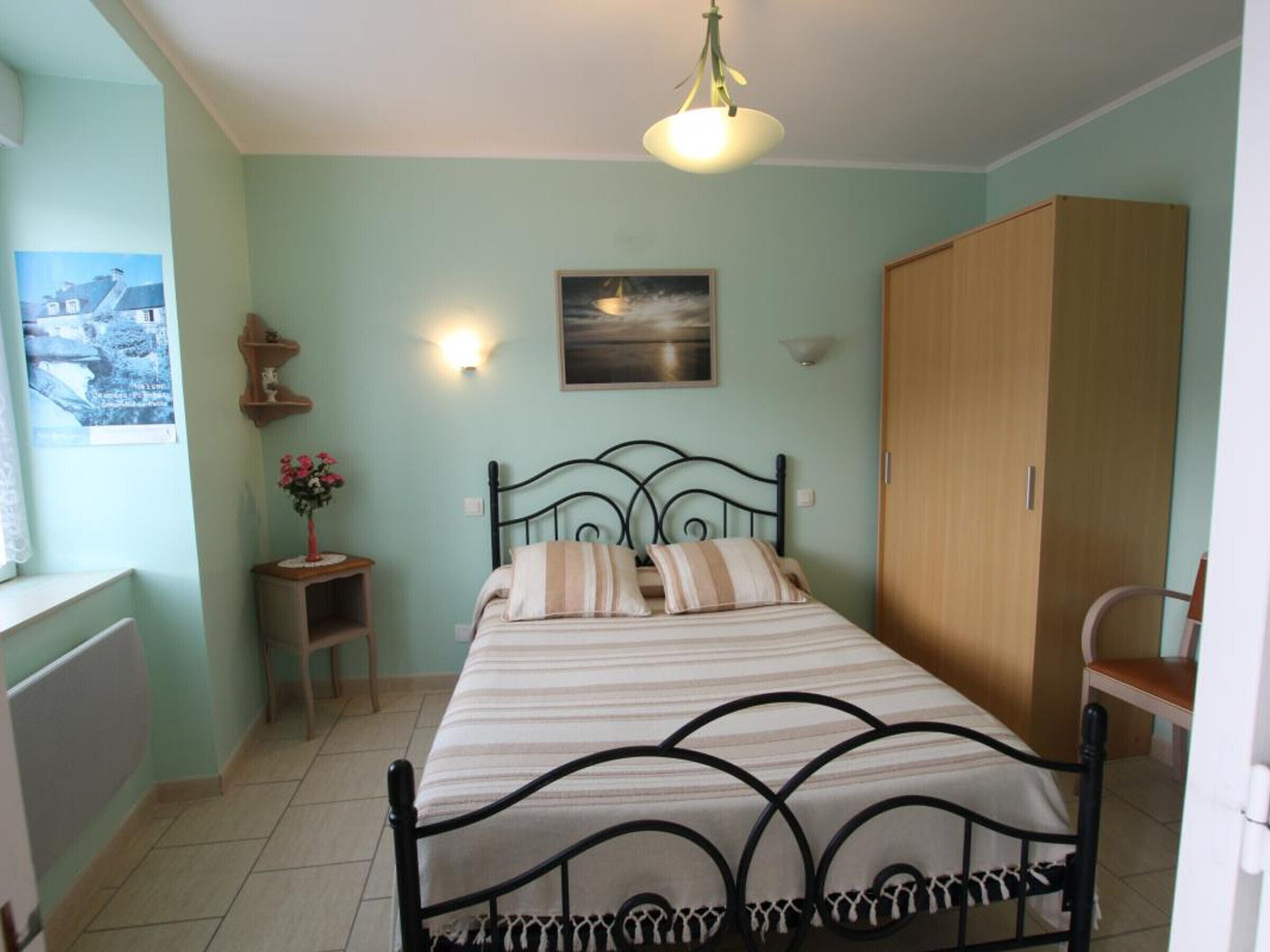 A bedroom with a bed, a wooden wardrobe, a small bedside table with a vase of flowers, a wall-mounted light, and a painting above the bed.
