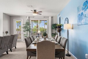 A dining room with a table set for six, chairs, a ceiling fan, and a view of palm trees through the windows.