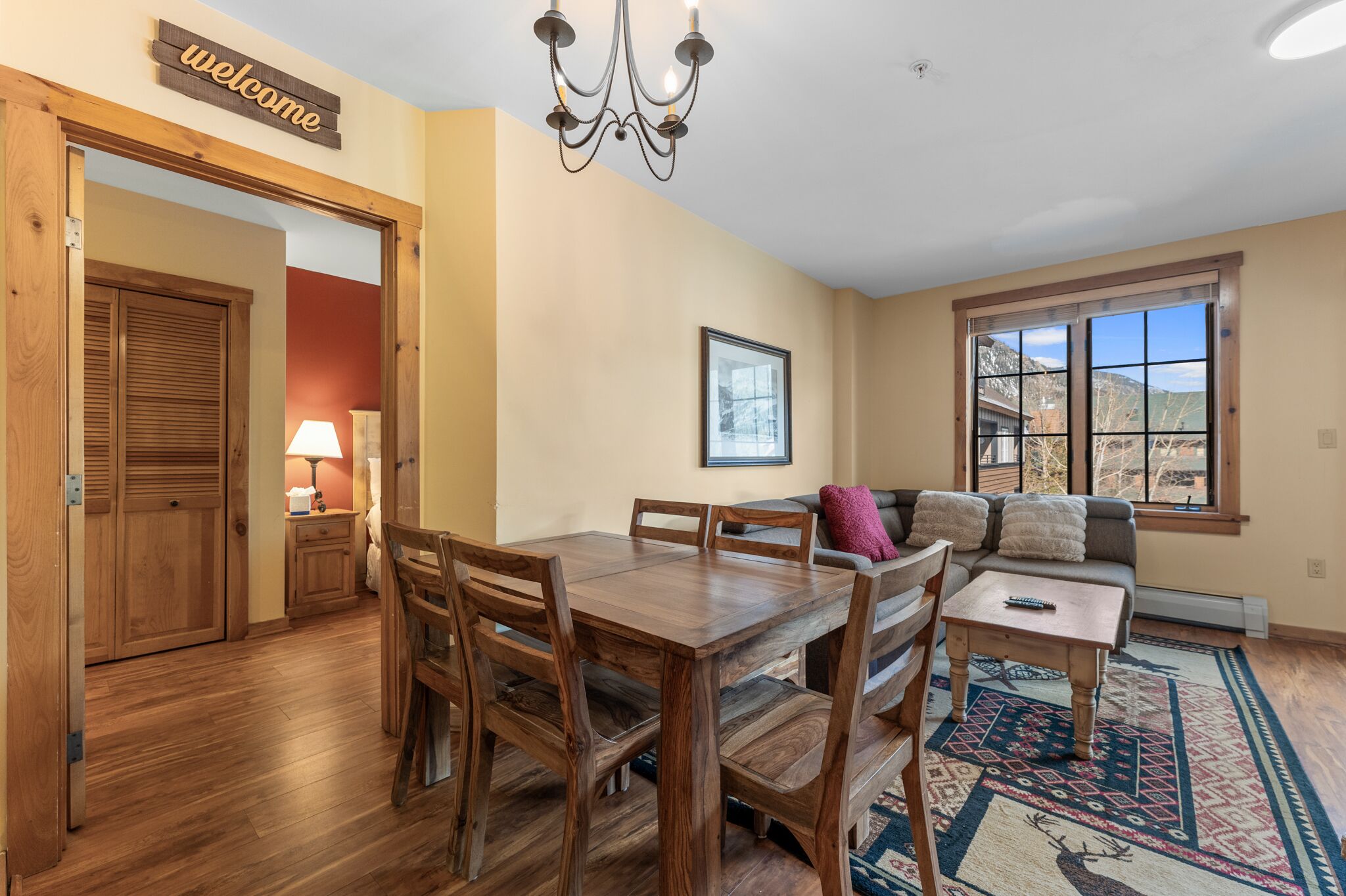 A dining area with a wooden table and chairs, a sofa, and a window with a mountain view.