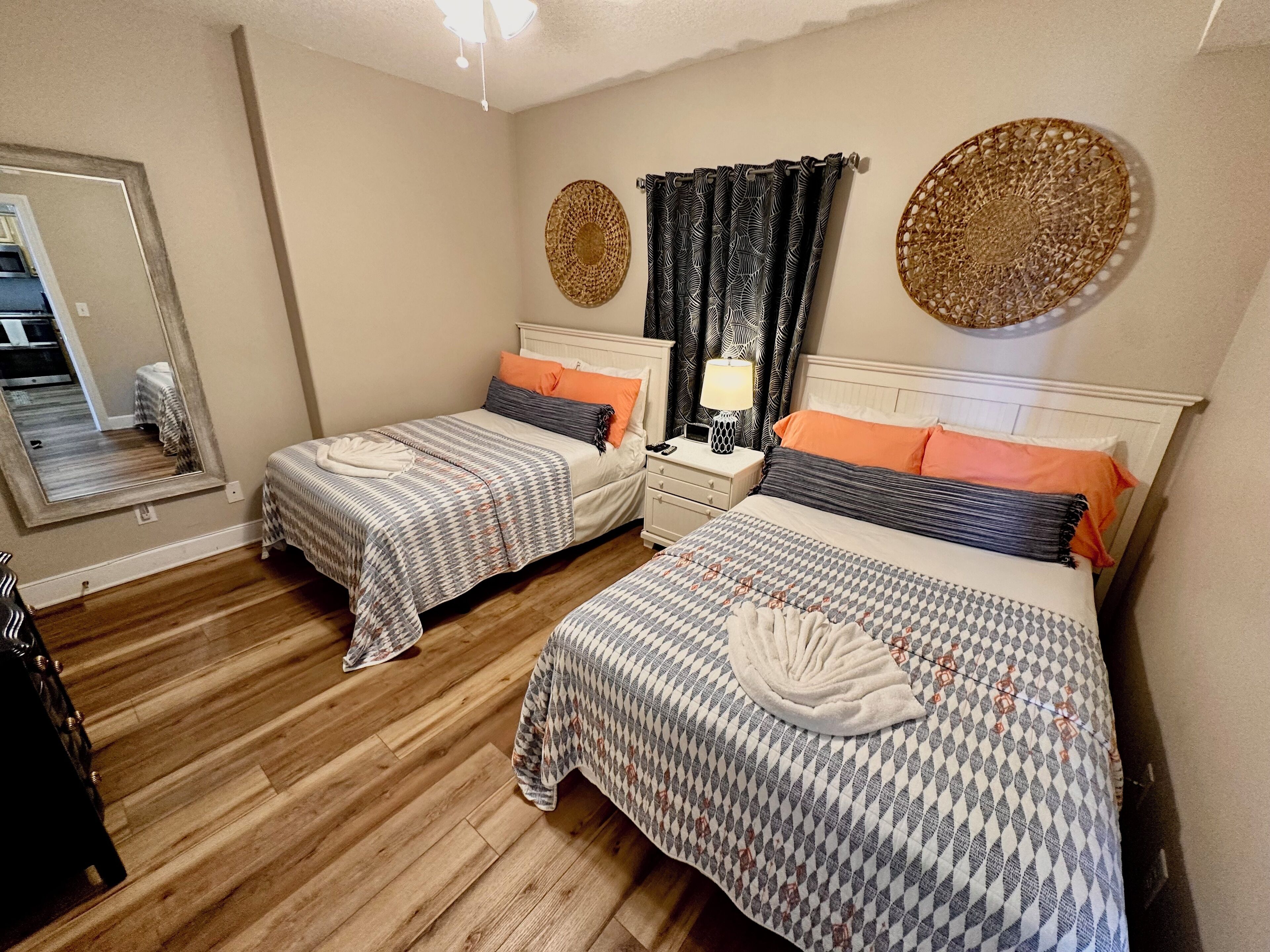 A bedroom with two beds, wooden flooring, a nightstand with a lamp, and decorative wall art.