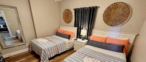 A bedroom with two beds, wooden flooring, a nightstand with a lamp, and decorative wall art.