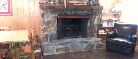 A cozy room with a stone fireplace, a wooden table, a leather chair, and a bookshelf.