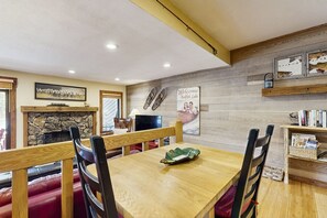 A dining area with wooden tables and chairs, a fireplace, and a wall-mounted shelf with books and a welcome sign.