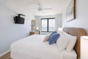 Habitación de hotel con cama, ventilador de techo, televisión, ventana con vistas, mesita de noche con lámpara y un cuadro en la pared.