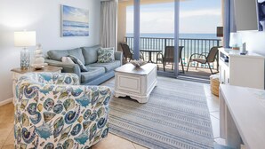 A modern living room with a sofa, armchair, and coffee table, a balcony with a view of the sea, and a dining area with chairs. - 2 BR @ Island Winds West - *Beach Front* (Gulf Shores)