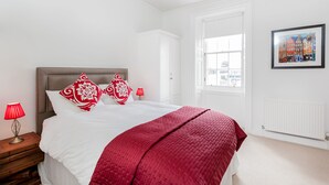 Ein Schlafzimmer mit einem Bett, einem Nachttisch, einer roten Lampe, einem weißen Heizkörper und einem Fenster mit Blick auf Gebäude.