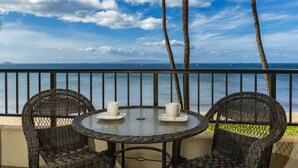 A balcony with wicker chairs and a table, overlooking a beach with palm trees and a clear sky.