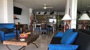 Ein Wohnzimmer mit Sofa, Sessel und Couchtisch. Es gibt eine Küchenzeile mit Schränken und einem Esstisch.