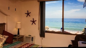 Ein Zimmer mit einem Bett, einem Nachttisch, einer Lampe und einem Fenster mit Blick auf Strand und Meer.