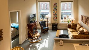 A compact living room with a sofa, rocking chair, and a TV. A window offers a view of buildings outside.