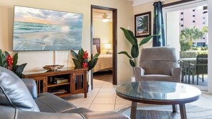 A living room with a leather sofa, a glass coffee table, a TV displaying a beach scene, a wooden cabinet, and a large window with a view of a building and palm trees. - Fun, Easy & Sunset Views! Ground Floor Escape – Islander 114 (Destin)