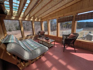 Une cabane en bois avec un grand lit, deux chaises, une petite table et de grandes fenêtres donnant sur une forêt.