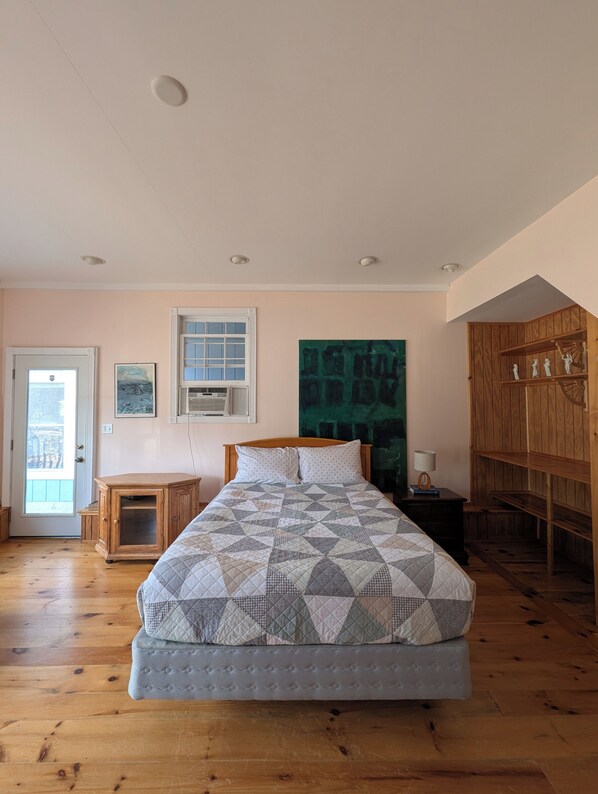 A bedroom with a bed, wooden floor, a door, a window, and a large painting.