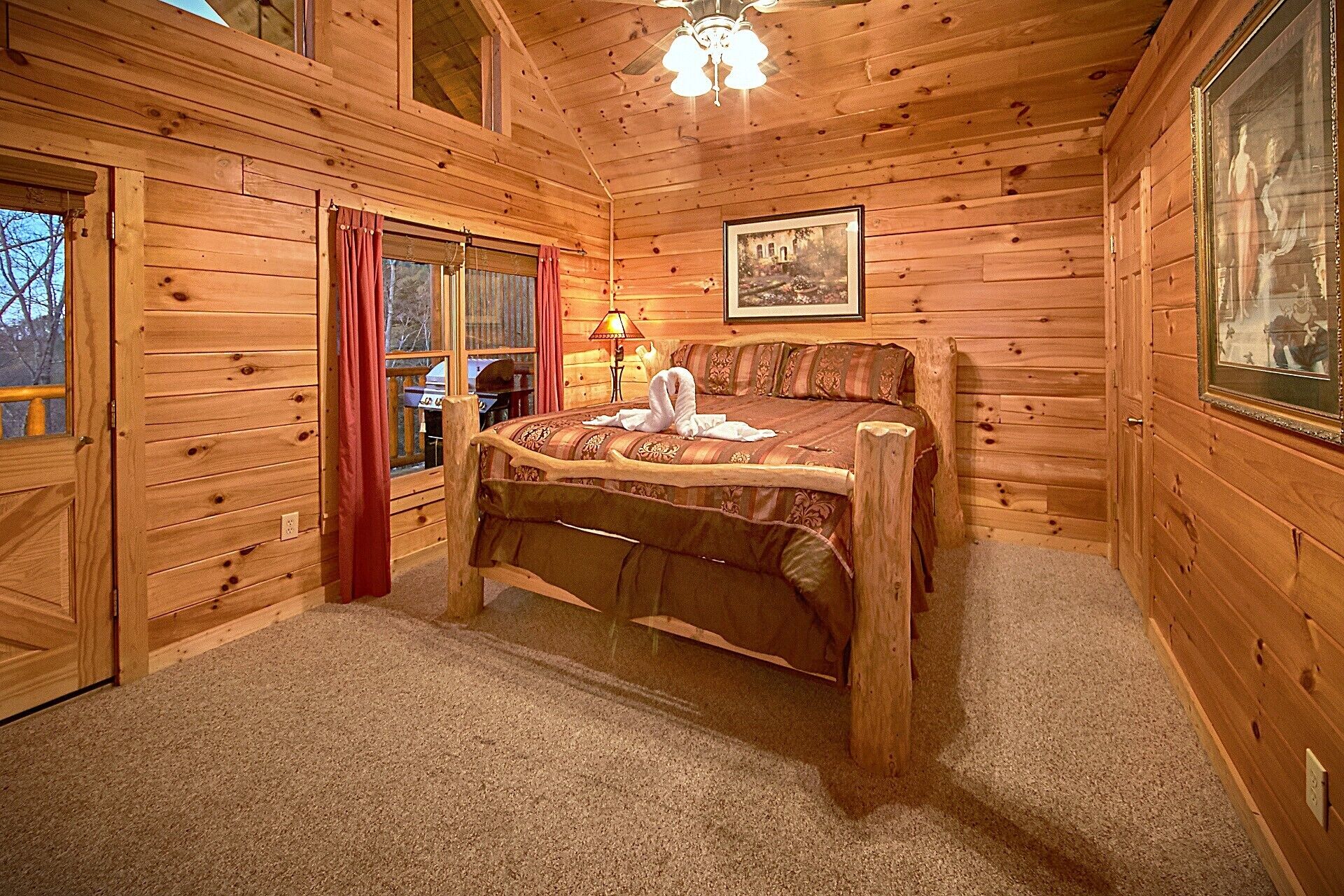 A wooden cabin bedroom with a bed, a lamp, a painting, and a window with curtains.