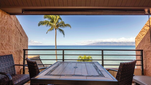 Un balcon avec une table en bois et des chaises en osier, donnant sur une plage avec un palmier et une vue sur l’océan.