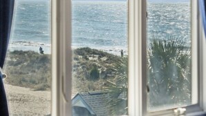 Een raam met uitzicht op zee en strand, gordijnen, en een deel van een bed.