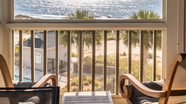 Een houten terras met twee stoelen en een tafel, met uitzicht op een strand met palmbomen en een hek.