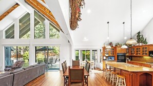 Un vaste espace de vie comprenant un grand canapé, une table à manger et des chaises, une cuisine dotée de meubles en bois et d’un îlot central, ainsi que de grandes fenêtres offrant une vue sur l’extérieur.
