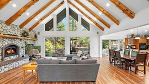 Un salon spacieux avec une cheminée en pierre, un grand canapé gris, des poutres en bois et une vue sur l’extérieur.