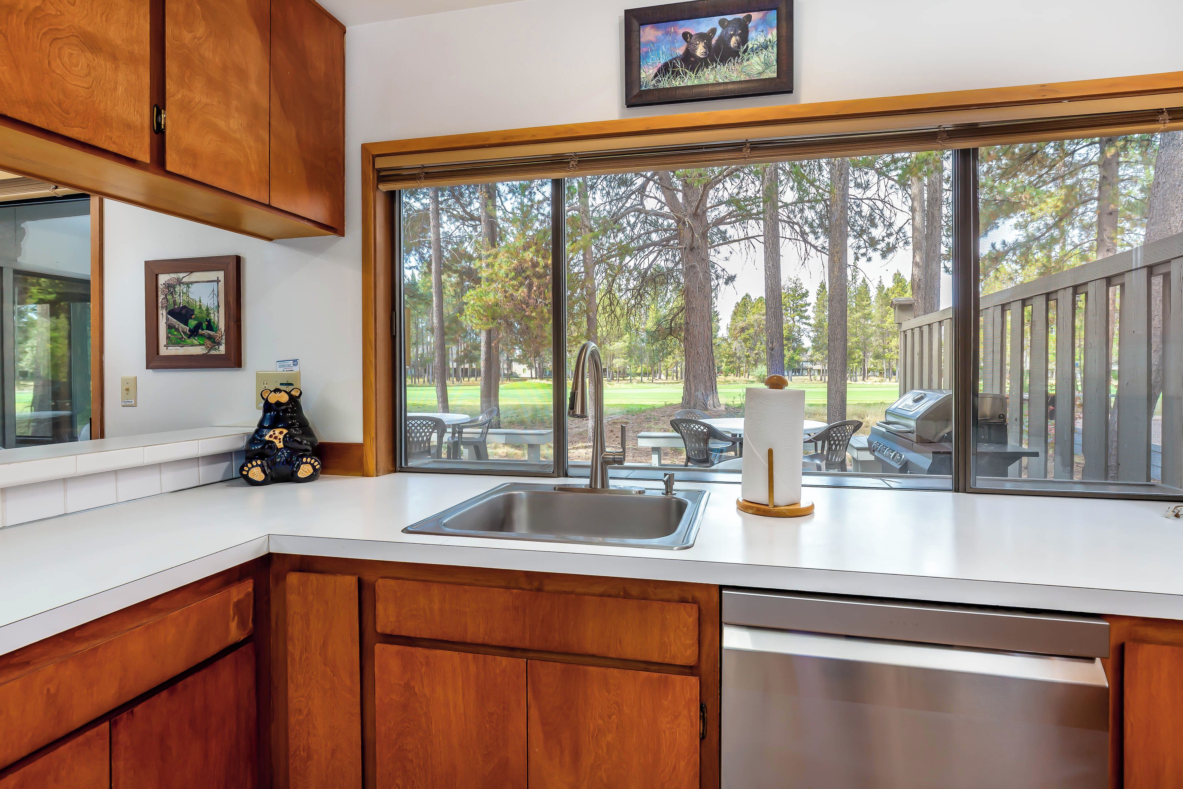 A kitchen with wooden cabinets, a stainless steel sink, and a large window overlooking a backyard with a patio and trees.