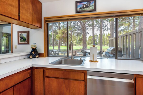 A kitchen with wooden cabinets, a stainless steel sink, and a large window overlooking a backyard with a patio and trees.