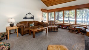 Ein gemütliches Wohnzimmer mit einem braunen Ledersofa, einem Holztisch, einem karierten Sessel und einem Schaukelstuhl. Große Fenster bieten Blick auf Bäume und eine entfernte Landschaft.